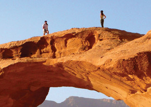 Two people stand atop a desert rock that arches like a bridge.