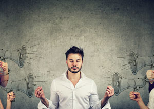 Young business man meditates while people shout at him with megaphones.