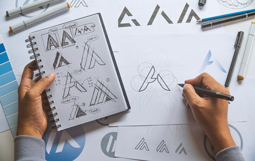 Person designing logo with a pen and paper. Holding journal with rough draft sketches for reference.